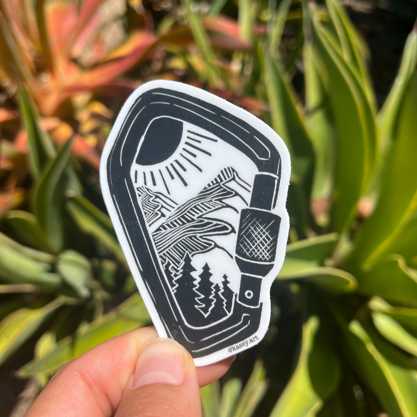 Black and white carabiner sticker with a design of a nature mountain scene against a green plant background