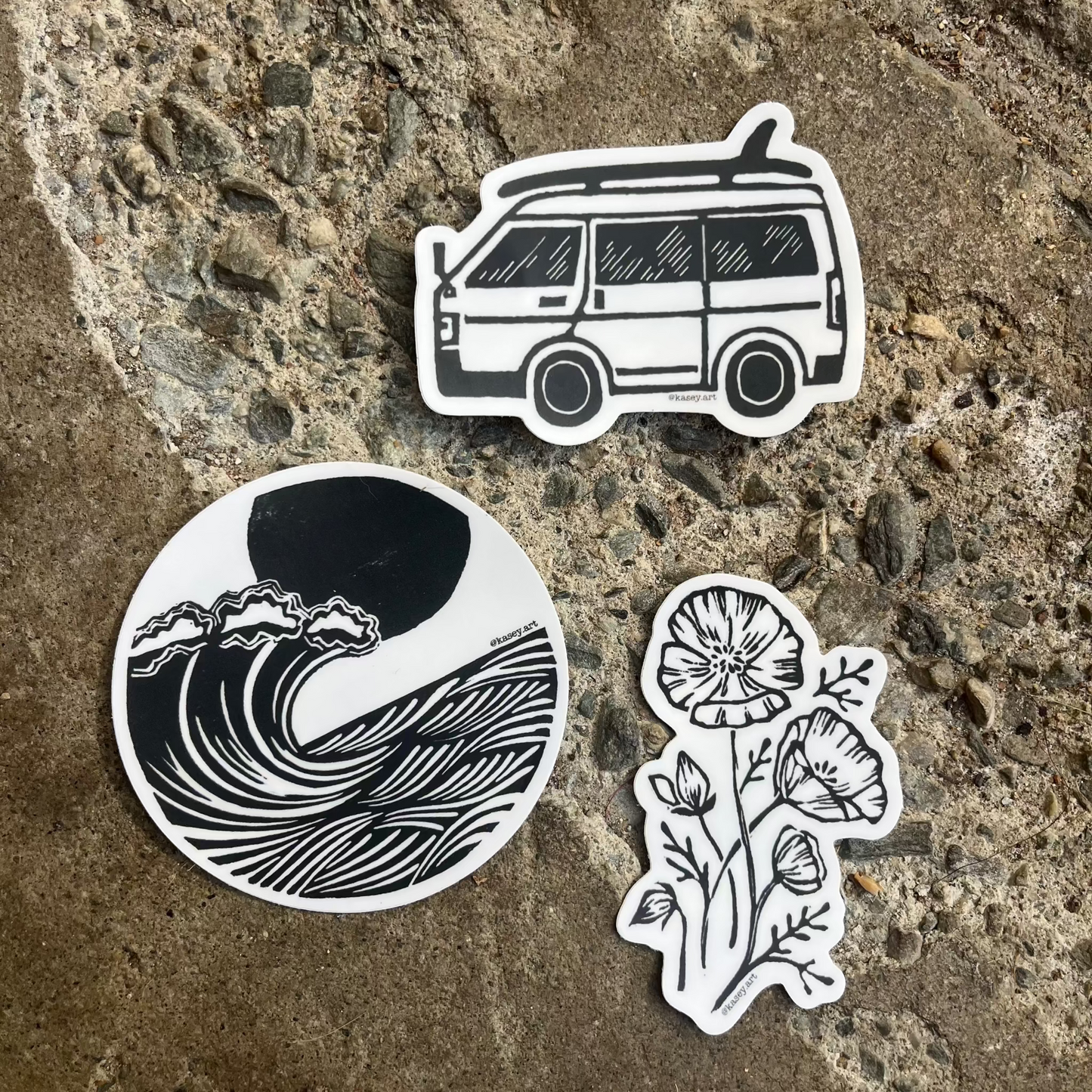 Three black and white stickers of a surf van, wave, and floral design on a textured stone surface.