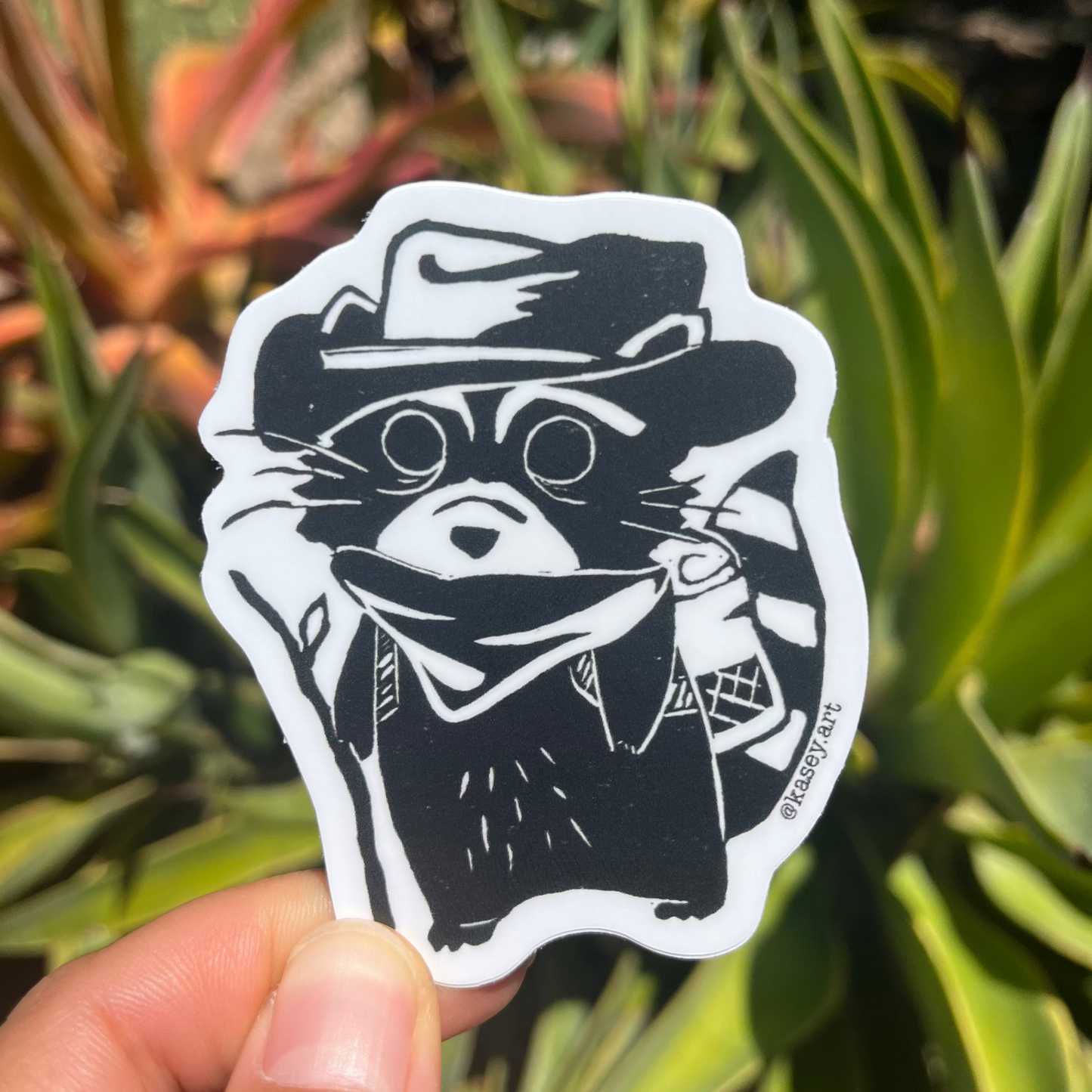 Black and white sticker of a stylized raccoon character with a cowboy hat in front of green plants.
