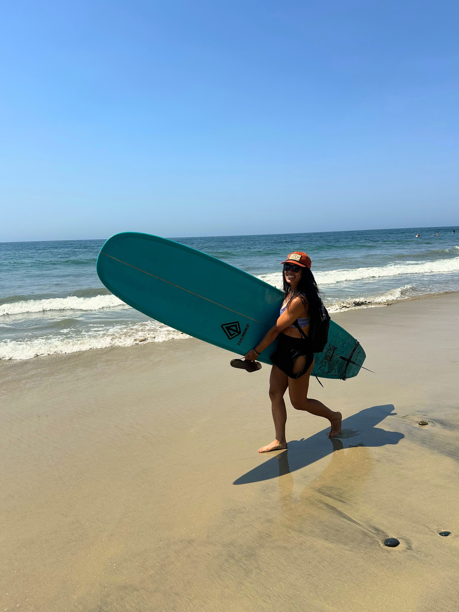 Person holding a teal surfboard on a sandy beach with ocean waves in the background