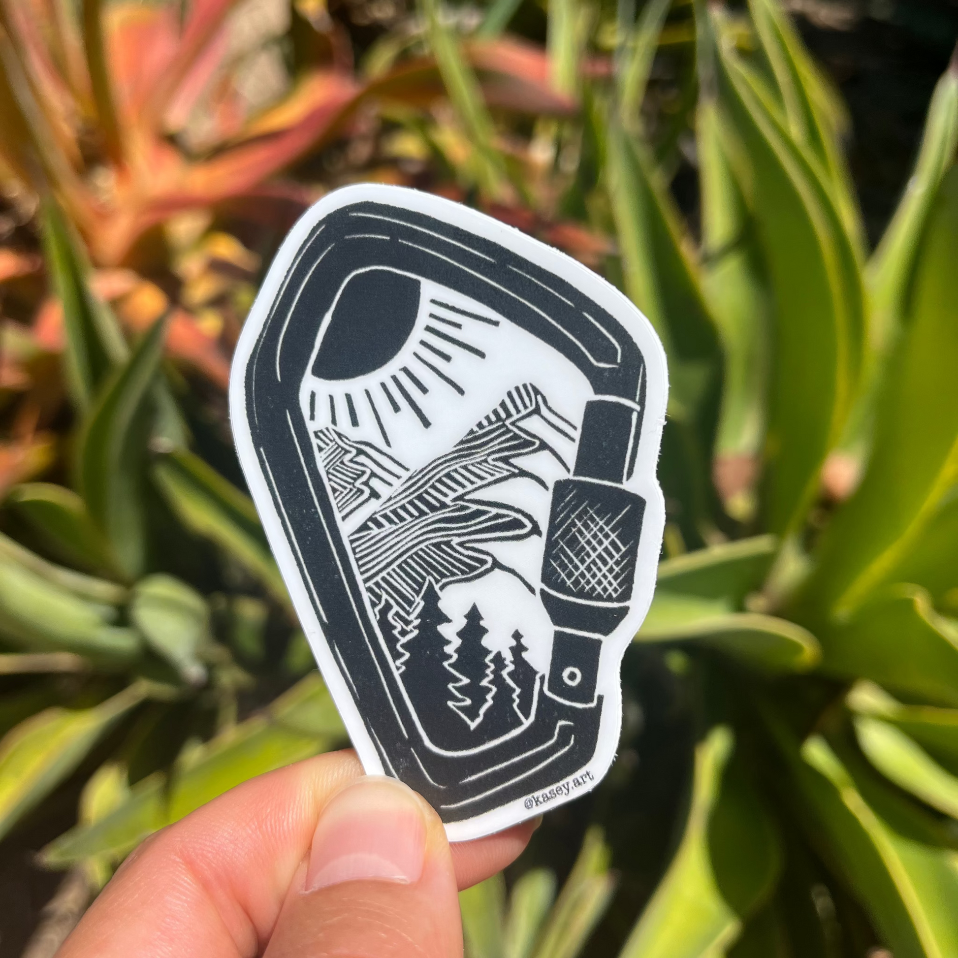 Black and white carabiner sticker with a design of a nature mountain scene against a green plant background