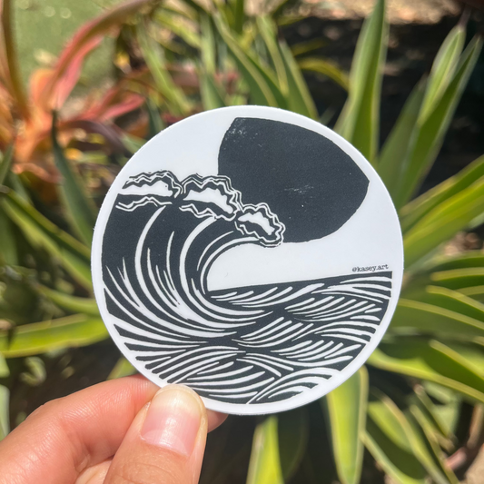 Round black and white sticker with ocean wave design held in front of green plants