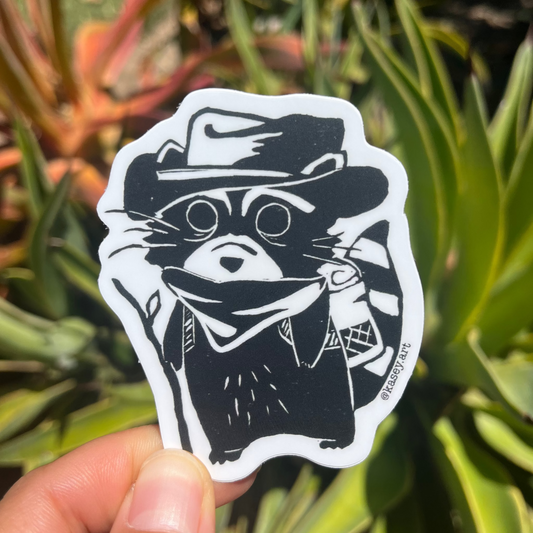 Black and white sticker of a stylized raccoon character with a cowboy hat in front of green plants.